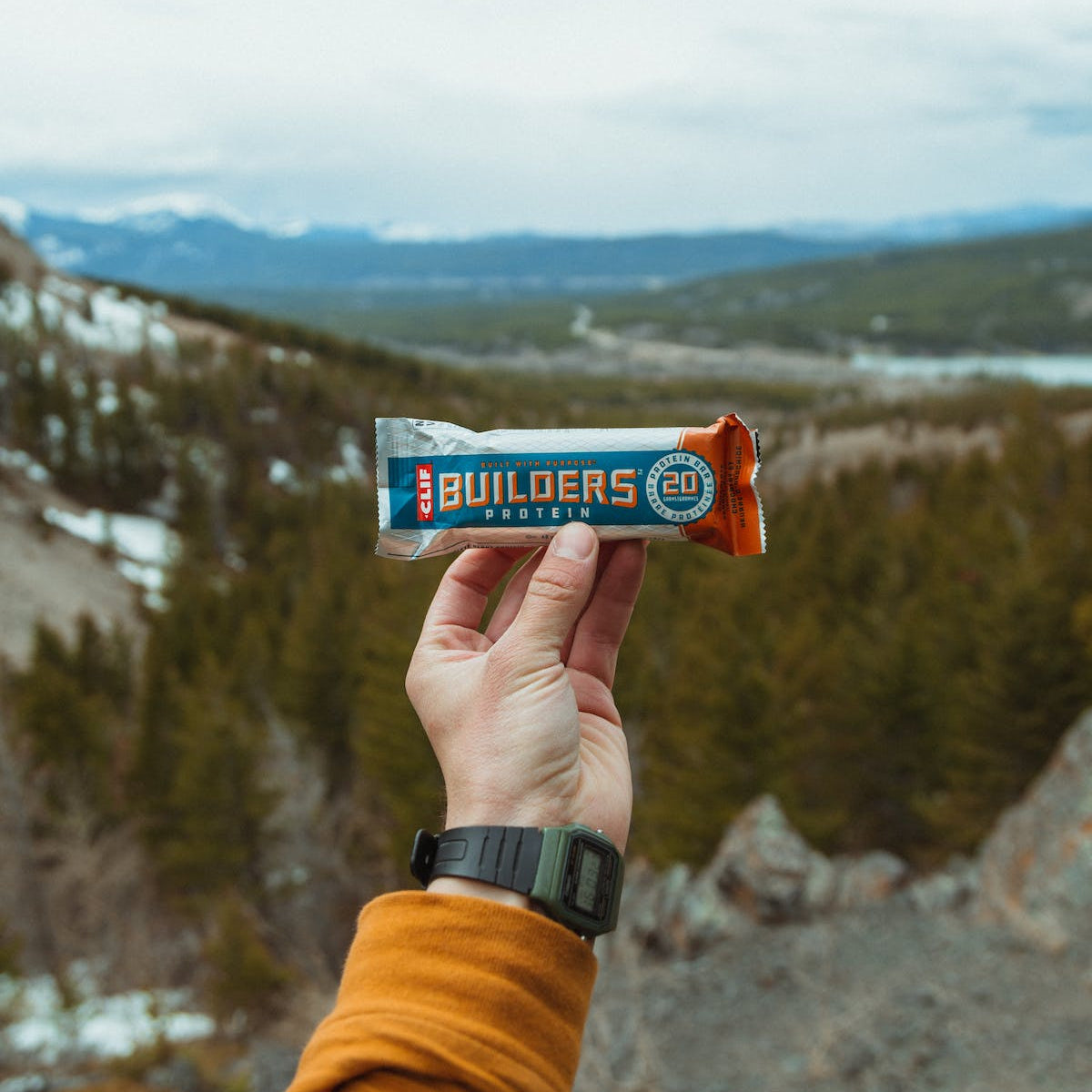 Someone holding Builder's protein bar on hike with mountains in back