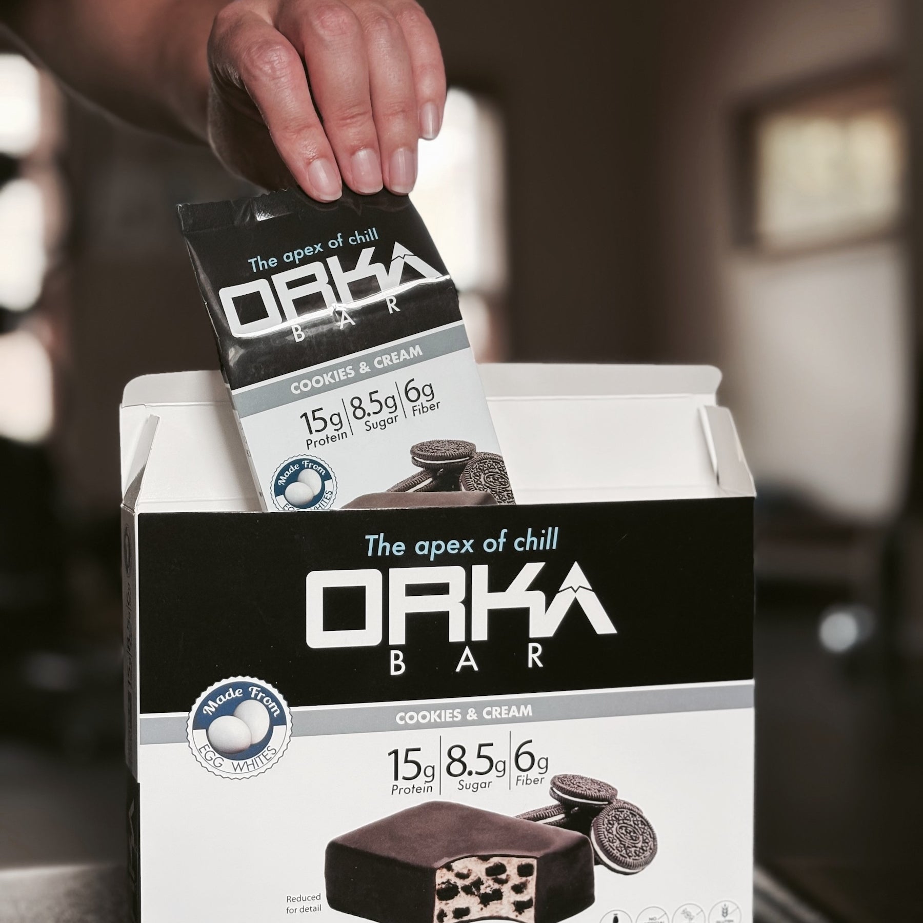 Orka Bar packaging with a hand holding a single bar above it, set against a blurred indoor background.