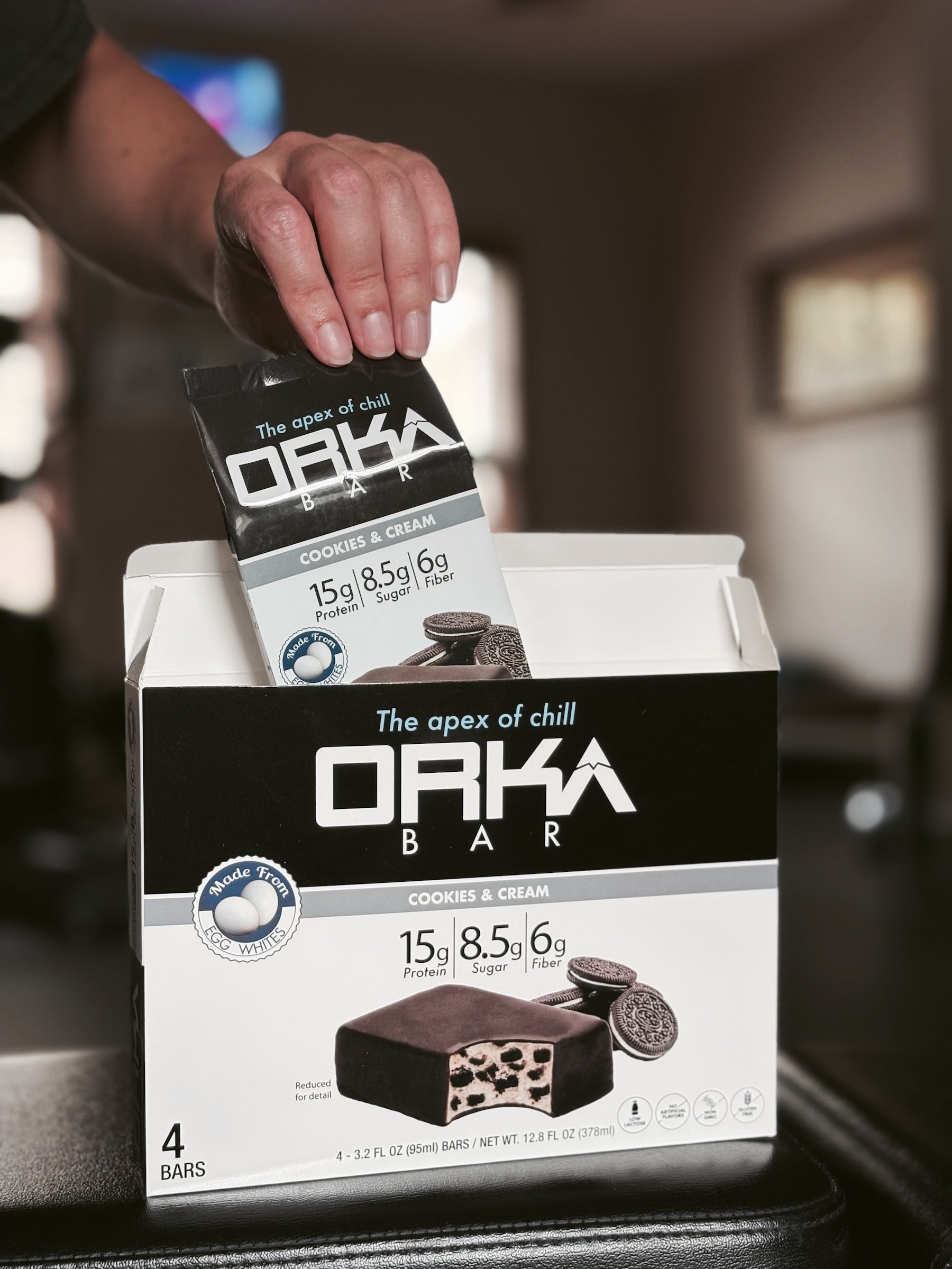 Orka Bar packaging with a hand holding a single bar above it, set against a blurred indoor background.